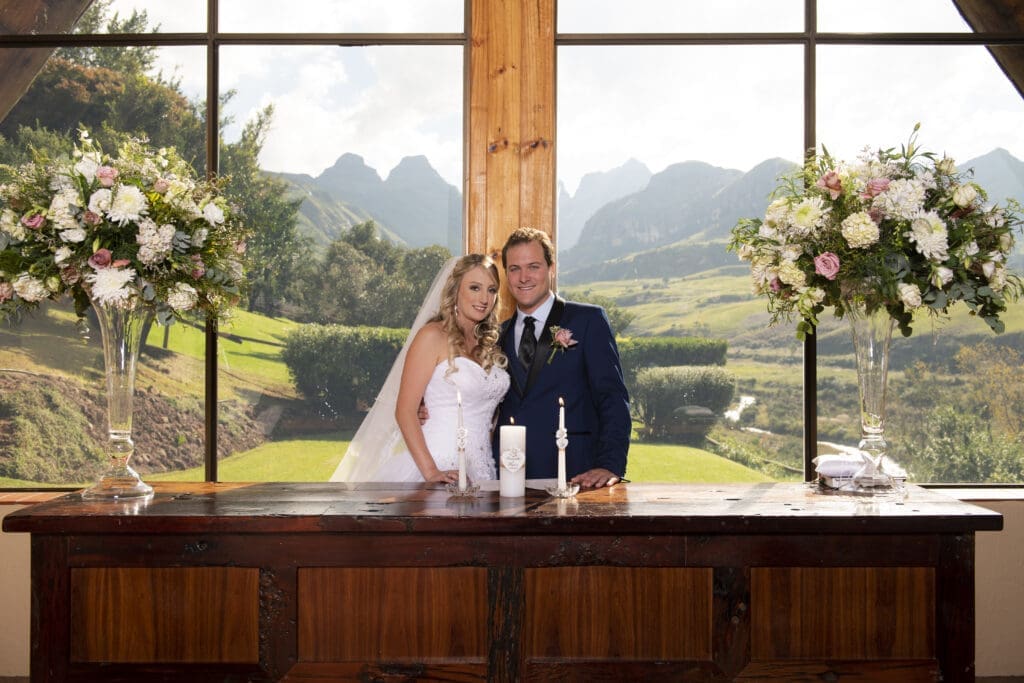 Bride and groom standing inside the chapel in front of wooden desk with glass windows and mountain peaks behind them.