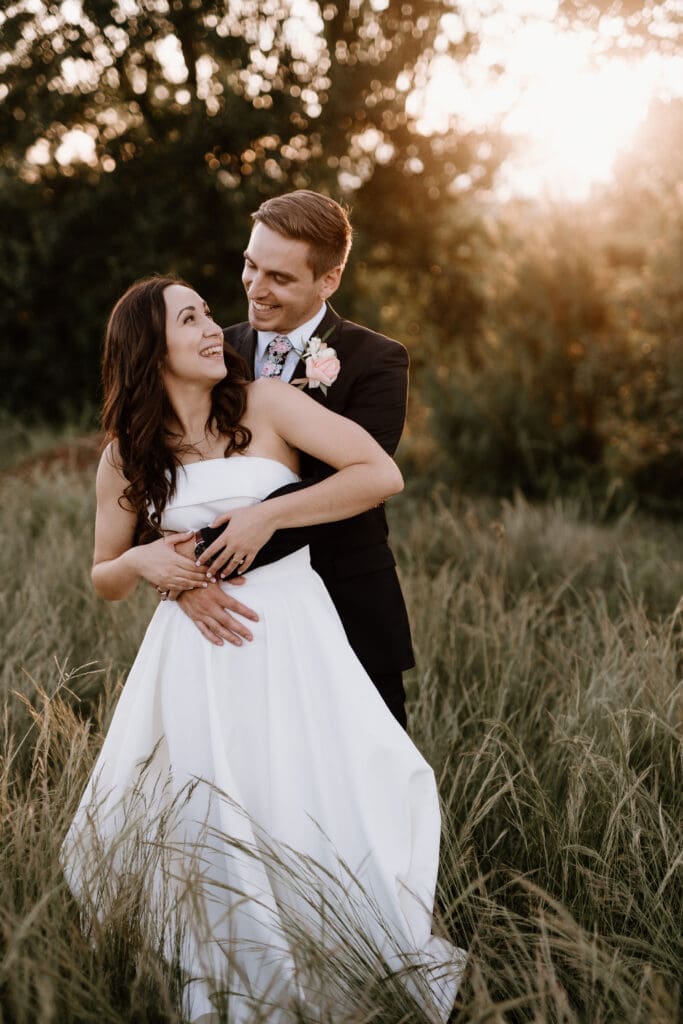 Bride and groom standing in long grass with sunset, bride looking back to groom