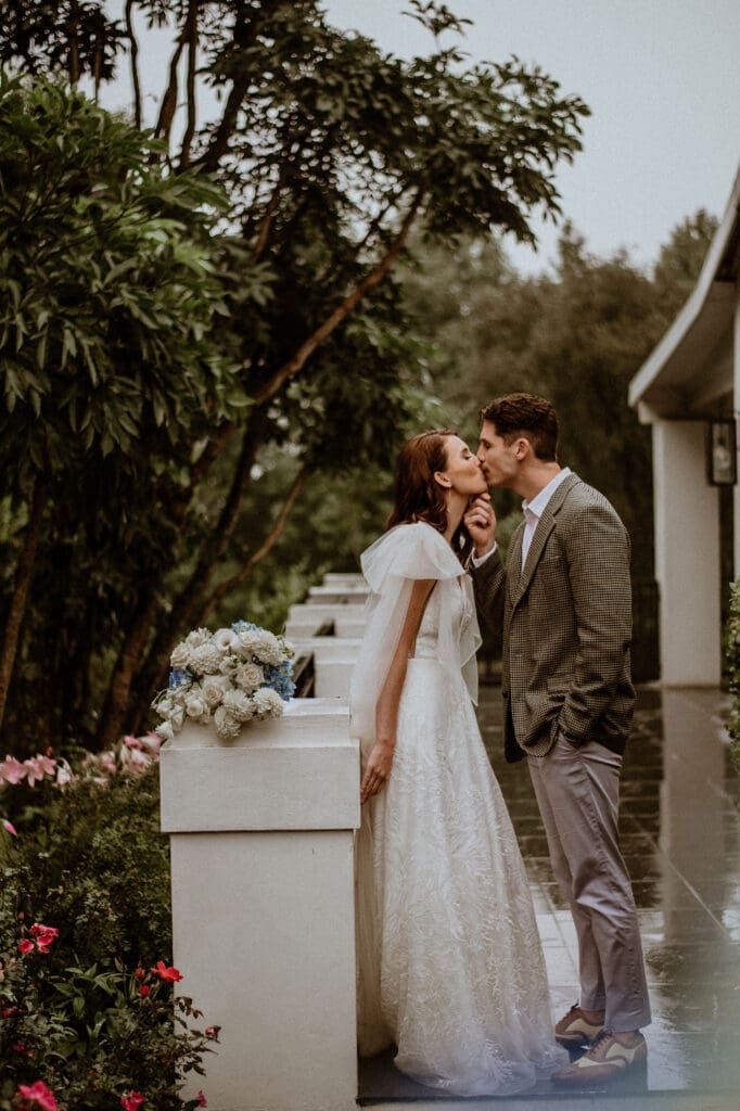 Bride leaning against low white wall and kissing groom.