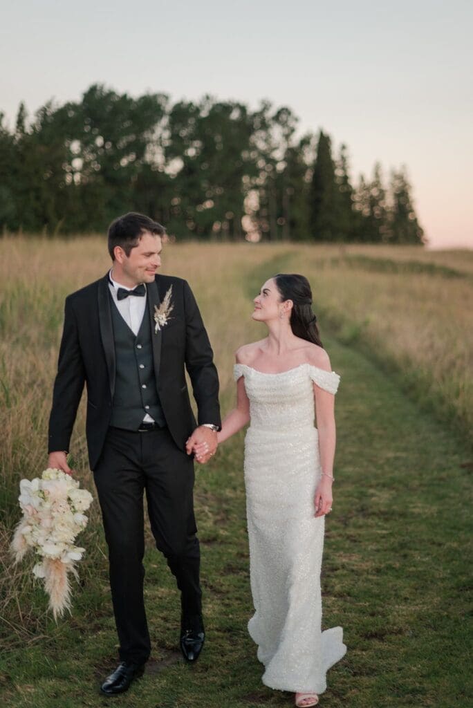 Bride and groom walking through grass field path, gazing at each other while groom carries bride's bouquet