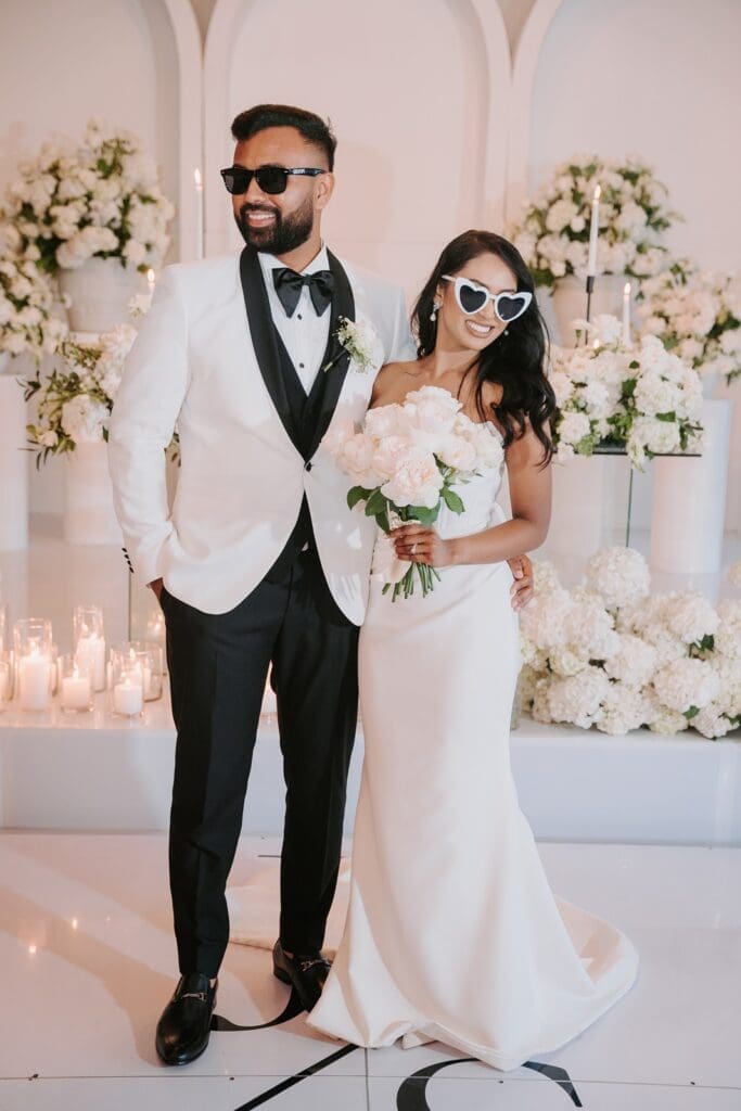 Bride and groom standing before floral display of white flowers wearing trendy sunglasses