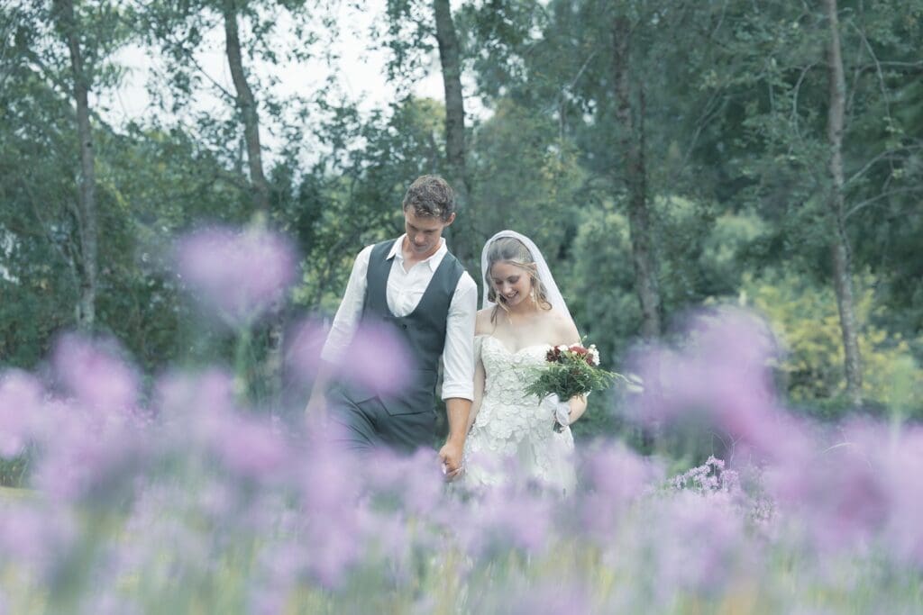 Bride and groom walking in field with hazy purple flowers in the foreground.